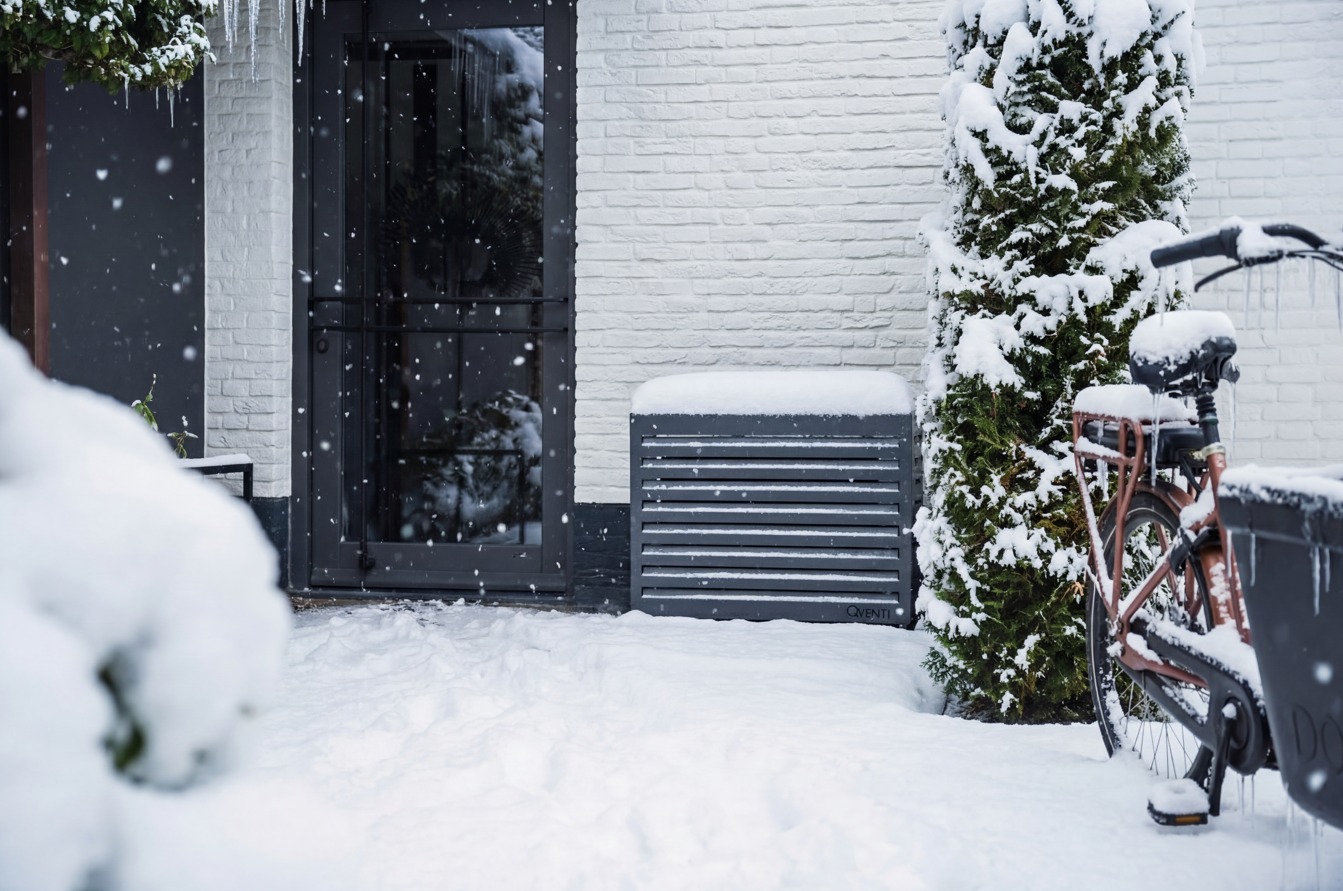 Moderne Qventi omkasting voor een warmtepomp of airco buitenunit, geplaatst tegen een witte gevel in een besneeuwde tuin, met zichtbare sneeuw op de omkasting, fiets en conifeer – toont winterbestendige installatie en esthetisch design.